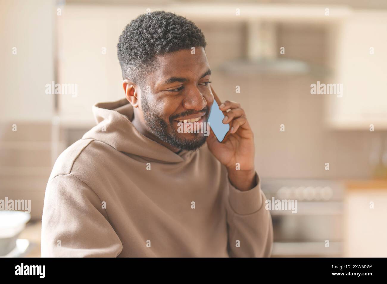 Homme heureux dans le sweat à capuche brun en utilisant le téléphone, étudier, apprendre, faire des achats, travailler en ligne, parler en étant assis sur un canapé à la maison Banque D'Images