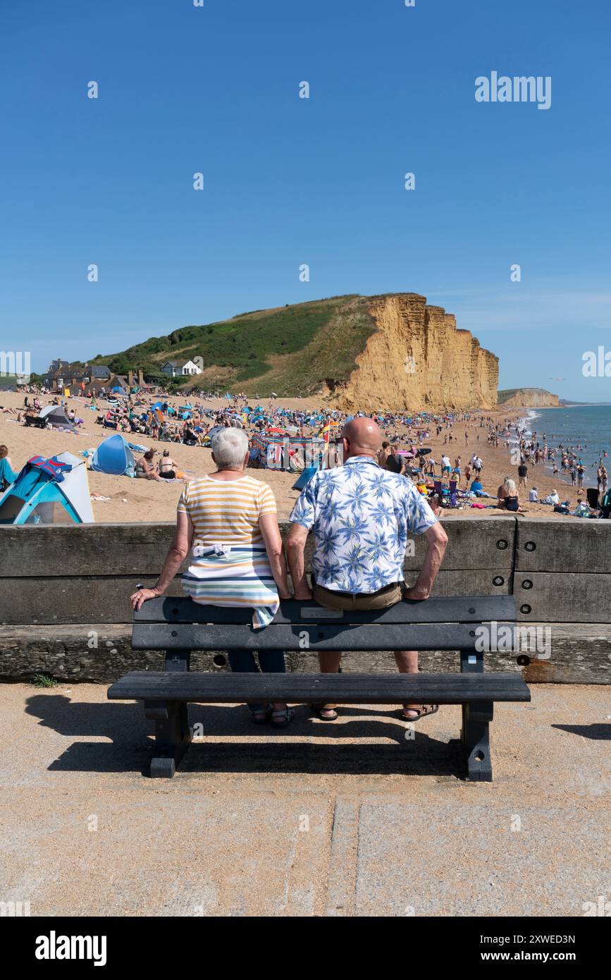Deux personnes sont assis sur un banc regardant une scène de plage animée par une chaude journée d'août à West Bay, au sud de Bridport, Dorset, Royaume-Uni. Banque D'Images