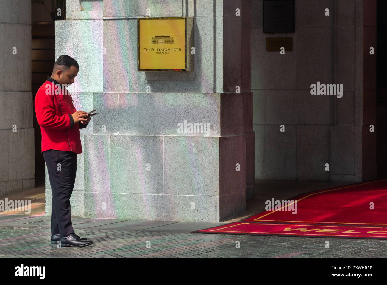 19 août 2024. Chasseur en uniforme rouge, regardant son téléphone portable, il est debout à l'entrée de l'hôtel Fullerton. Secteur des services. Singapour. Banque D'Images