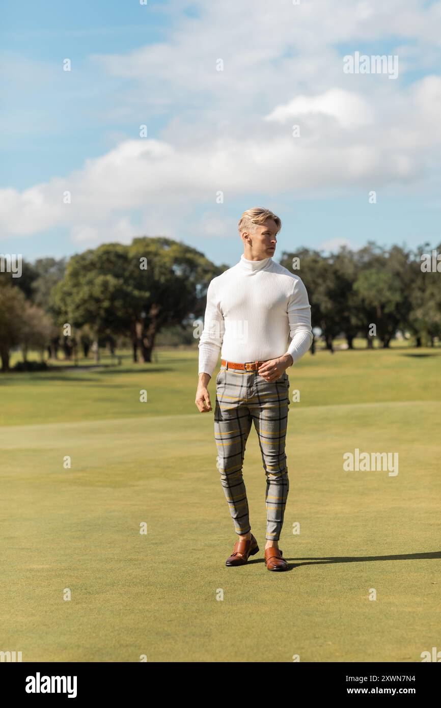 Un homme blond marche sur un terrain de golf vert à Miami, portant un pull blanc à col roulé et un pantalon à carreaux. Banque D'Images