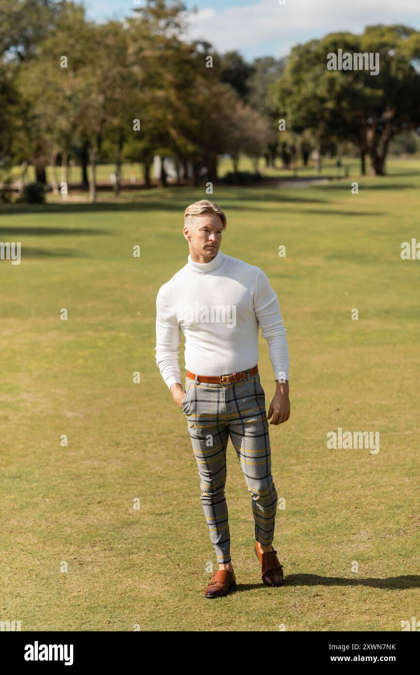 Un homme blond à col roulé blanc et pantalon à carreaux traverse un parc herbeux à Miami. Banque D'Images