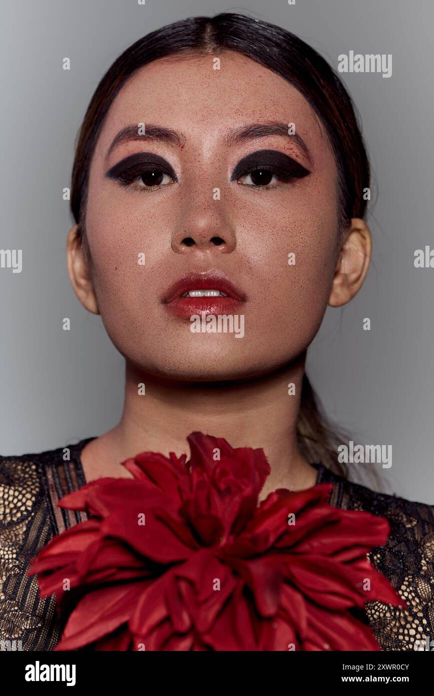 Femme asiatique avec le maquillage audacieux des yeux et une fleur pourpre regarde à la caméra. Banque D'Images