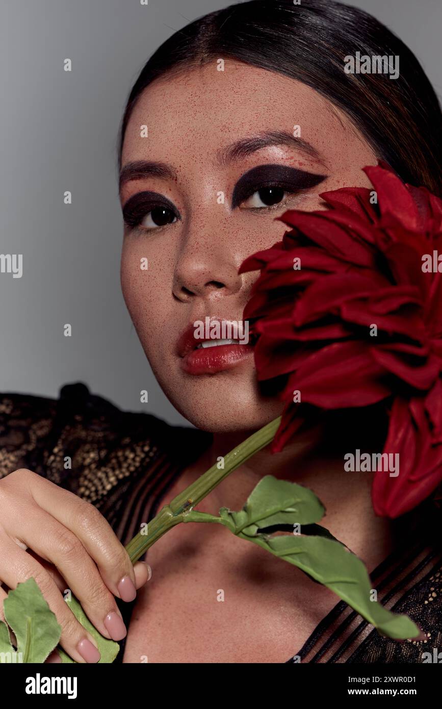 Une jeune femme aux cheveux foncés et au maquillage audacieux tient une fleur rouge contre son visage. Banque D'Images