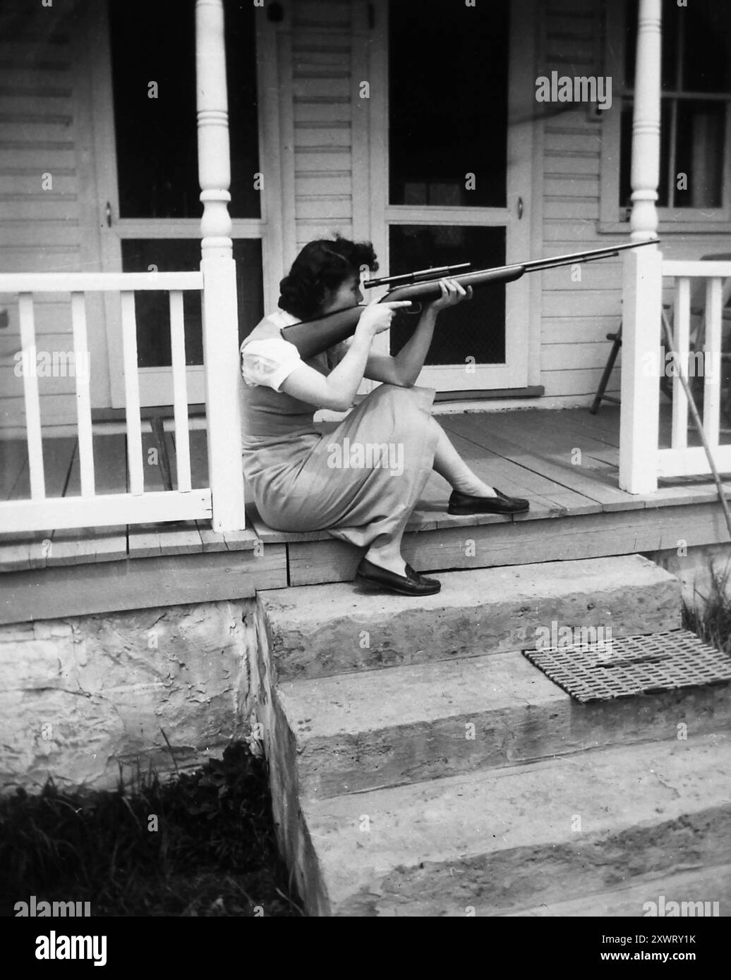 Jeune femme regarde son fusil tout en étant assise sur son porche, vers 1940. Banque D'Images