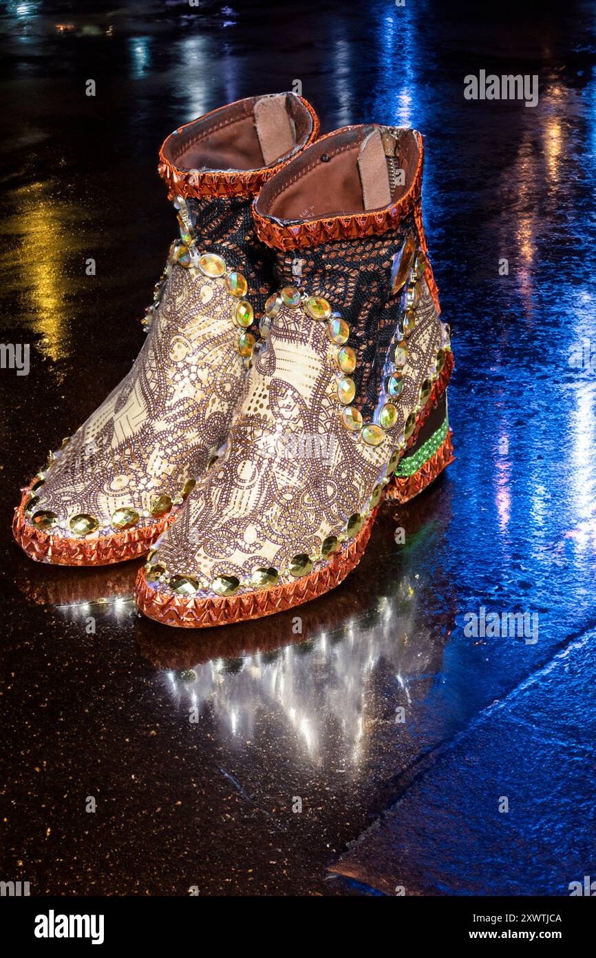 Une paire de bottes dorées au design complexe avec des embellissements colorés et des bijoux sur un trottoir humide, reflétant les néons la nuit. Banque D'Images