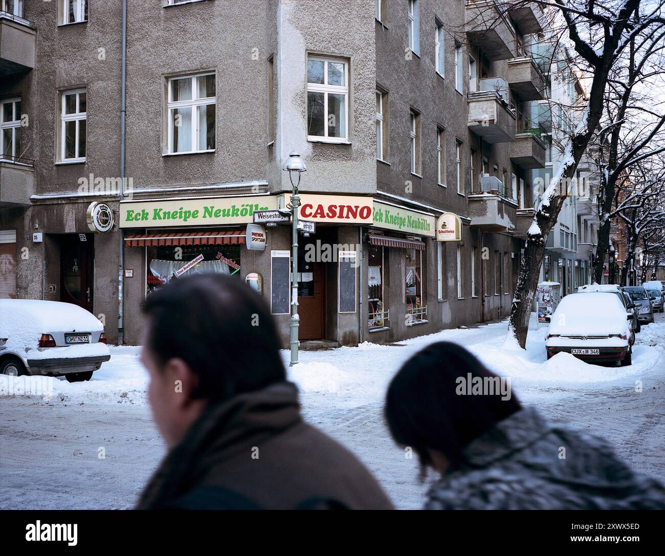 Une vue hivernale sur la rue de Berlin-Neukölln avec le pub d'angle Eck Kneipe Neukölln et un casino. Les trottoirs enneigés et deux piétons créent une atmosphère hivernale urbaine typique dans ce quartier allemand. Banque D'Images