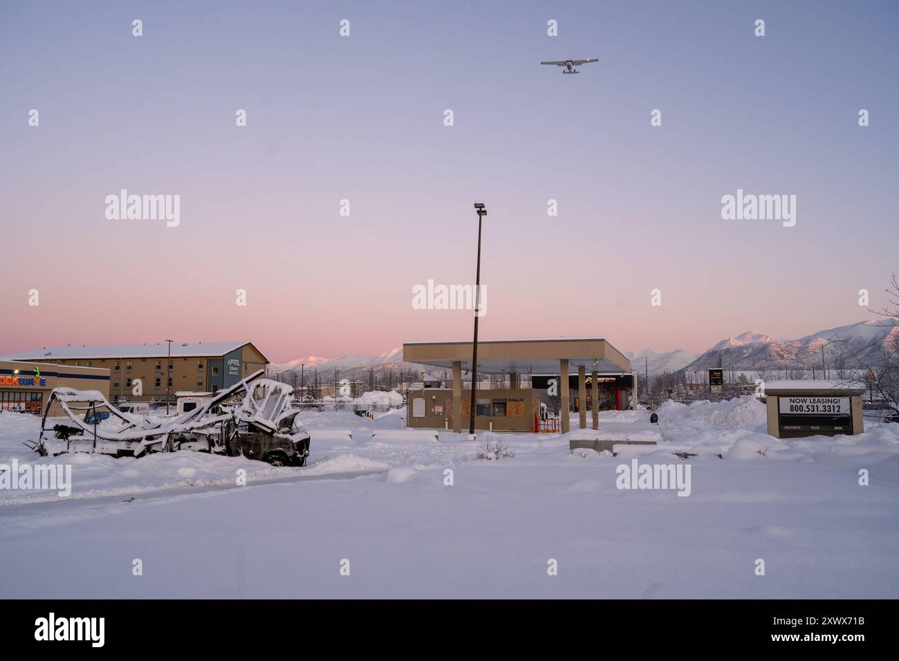 Un paysage hivernal enneigé près de l'aéroport de Merrill Field à Anchorage, Alaska. L'image montre un petit avion volant au-dessus de la tête, un parking couvert de neige et divers bâtiments sous un ciel pastel. Banque D'Images