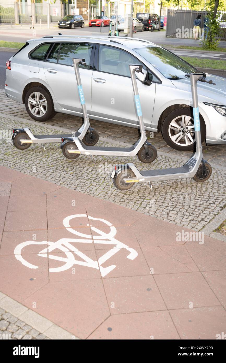 Trois scooters électriques sont garés sur une piste cyclable, bloquant la zone cyclable désignée, près d'une voiture garée. La scène se déroule dans une rue de la ville de Berlin, mettant en évidence les défis du transport urbain. Banque D'Images