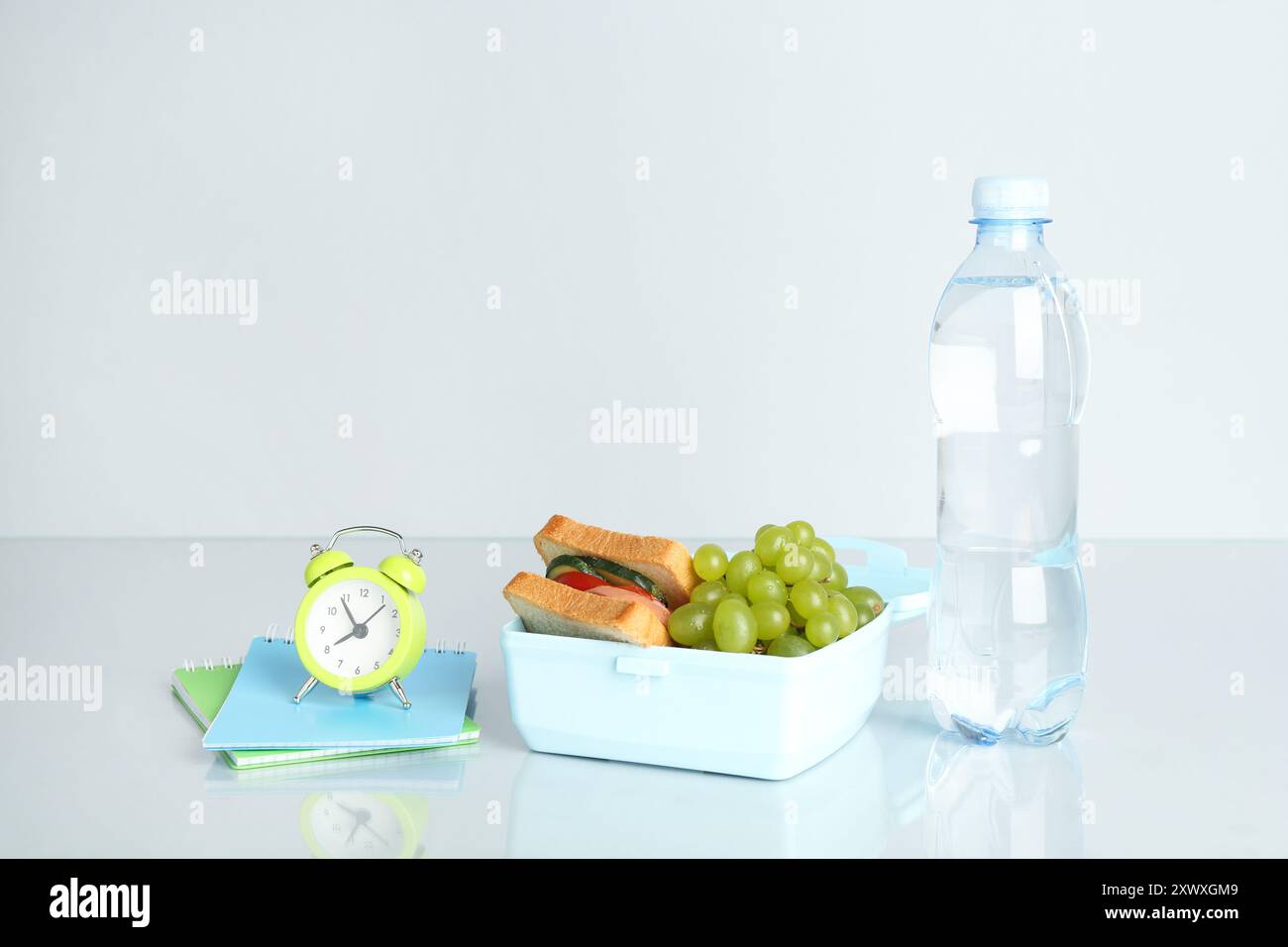 Boîte à lunch avec collations, bouteille d'eau, réveil et papeterie sur fond blanc Banque D'Images