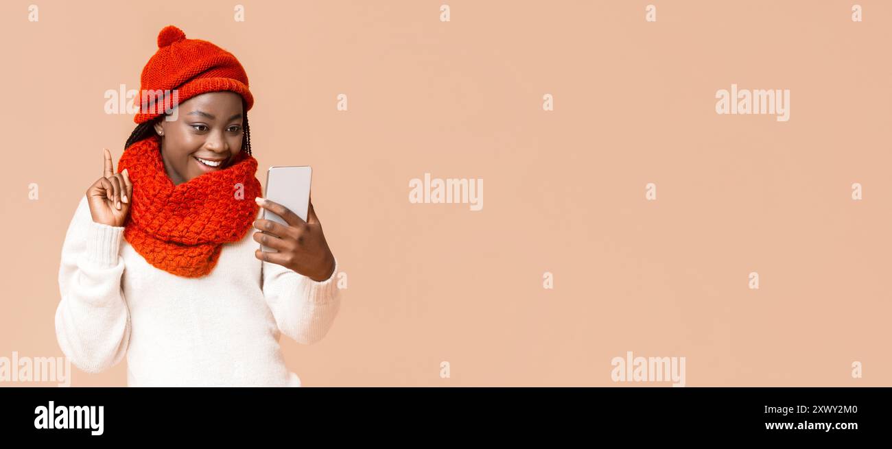 Joyeuse jeune femme dans une tenue d'hiver confortable s'engageant avec smartphone Banque D'Images