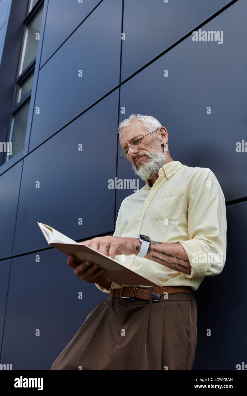 Un homme gay d'âge moyen avec une barbe lit un livre tout en se tenant devant un bâtiment moderne. Banque D'Images