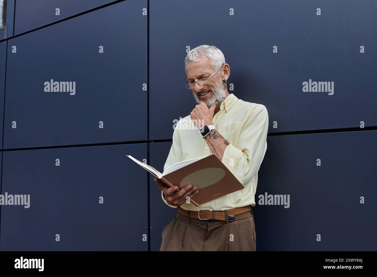 Un homme gay mûr avec une barbe lit un livre tout en se tenant debout à côté d'un bâtiment moderne, touchant soigneusement son menton. Banque D'Images