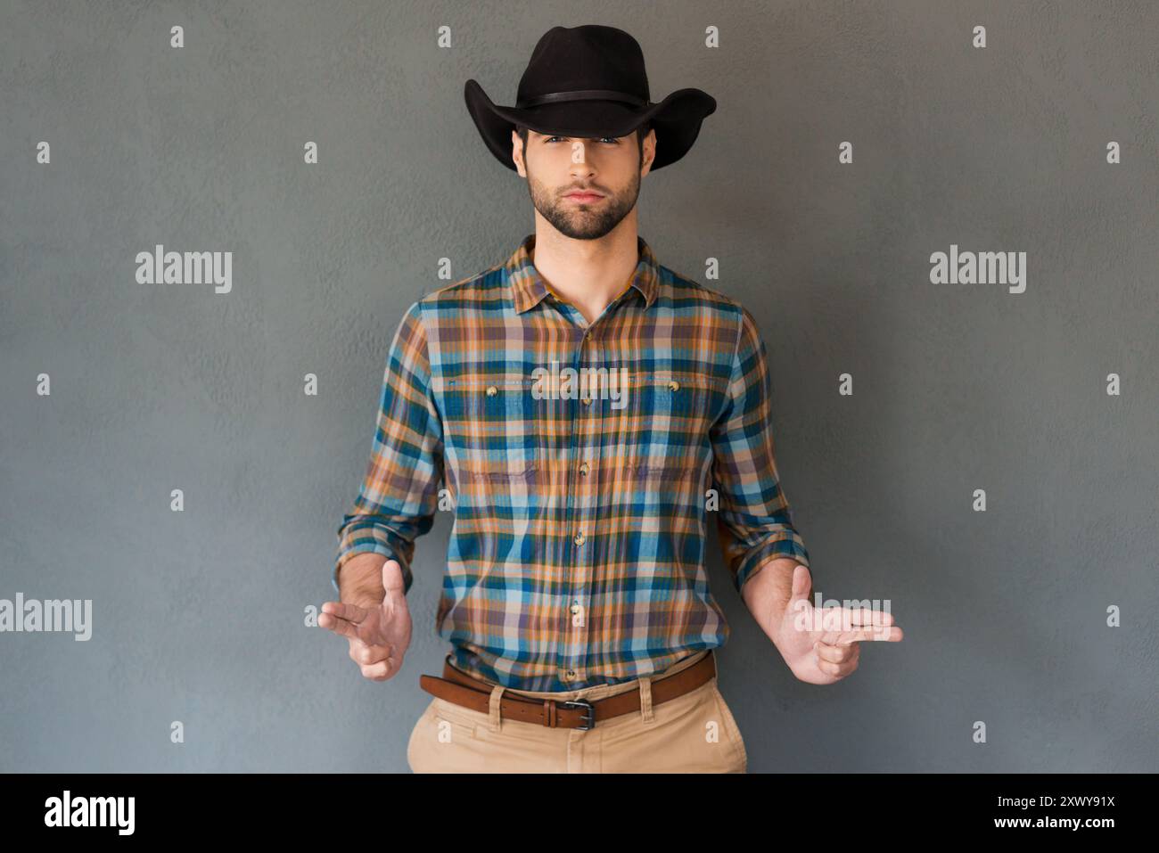 Mains levées ! Beau jeune homme portant un chapeau de cow-boy et faisant des gestes tout en se tenant debout sur fond gris Banque D'Images