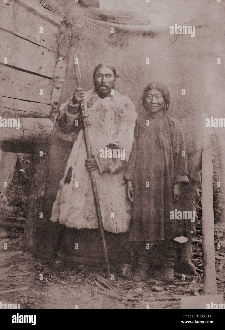 Peuples autochtones minoritaires du Nord, de Sibérie et de l'extrême-Orient de la Russie. Nivkh femme et homme debout près d'un mur de bois (groupe habitant la moitié nord de l'île Sakhaline). Un homme vêtu d'un long manteau de fourrure tient une lance dans ses mains. Une femme en vêtements simples sombres et de longues bottes. De l'expédition au Kamtchatka et aux îles commander par le célèbre naturaliste et médecin polonais. Benedykt Dybowski. Empire russe. années 1870 Banque D'Images