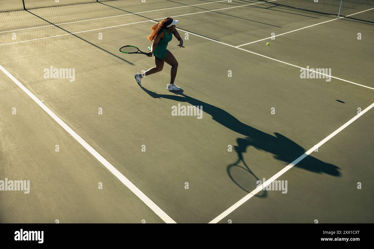 Femme jouant au tennis au coucher du soleil, balançant sa raquette et jetant une longue ombre sur le court Banque D'Images