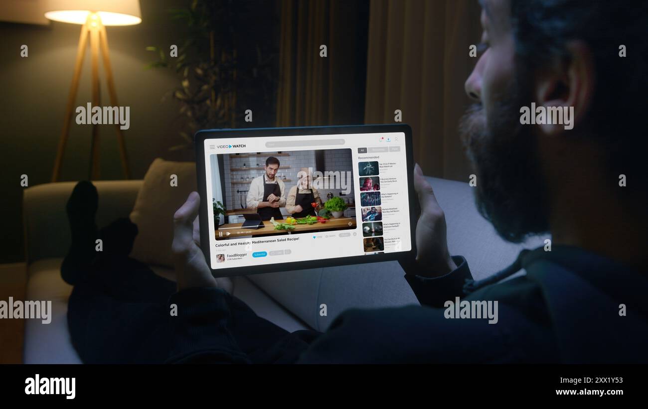 L'homme regarde un tutoriel de cuisine sur une tablette tout en étant allongé sur un canapé dans le salon. Apprentissage numérique moderne et commodité d'accès à l'éducation Banque D'Images