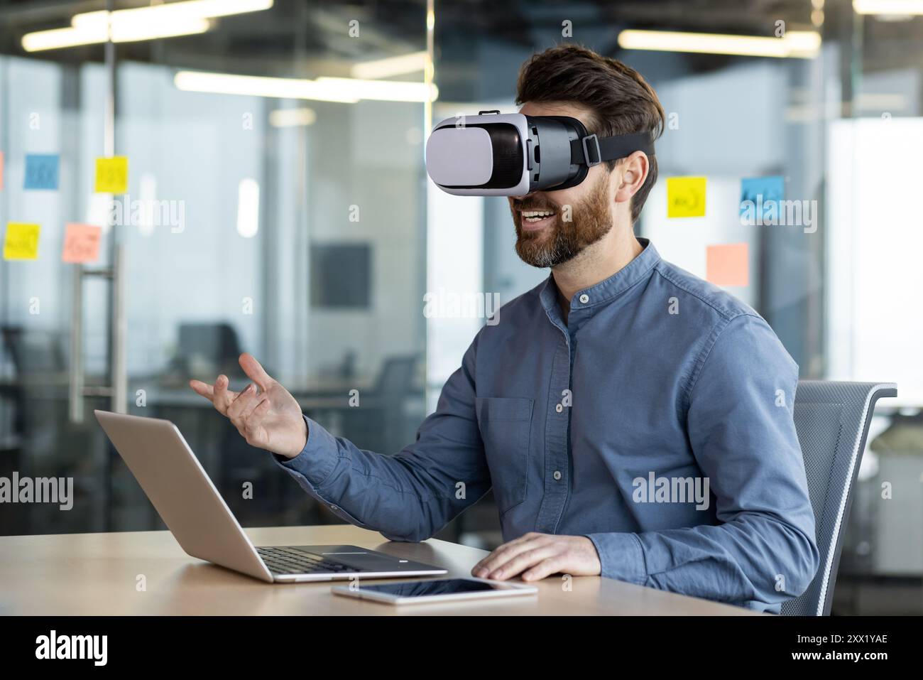 Un jeune homme d'affaires portant un masque virtuel parle à distance lors d'un appel vidéo, assis à un bureau dans le bureau et faisant des gestes avec ses mains. Banque D'Images