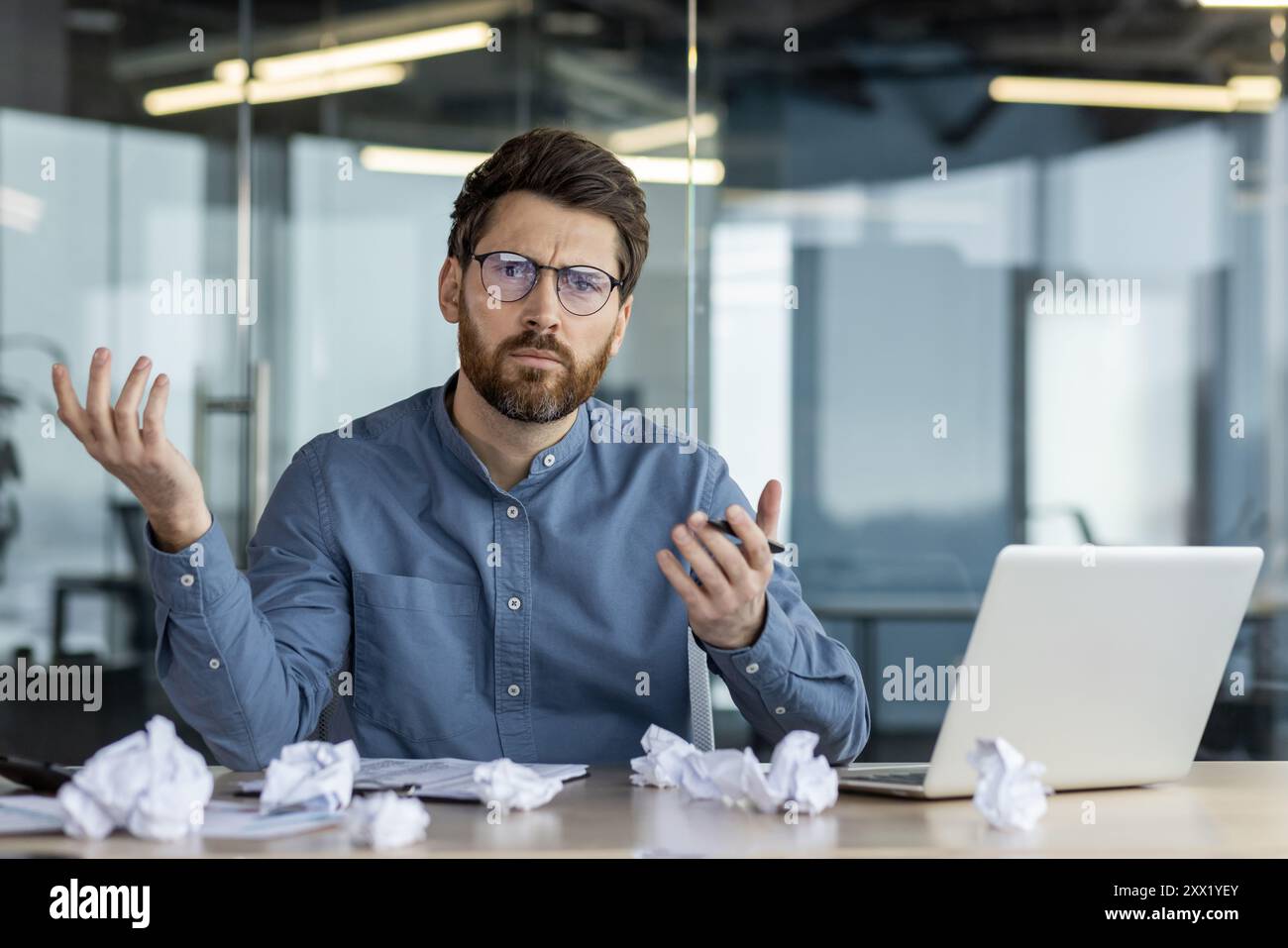 Portrait d'un jeune homme inquiet assis dans le bureau à un bureau avec du papier froissé dessus et regardant avec déception la caméra. Banque D'Images