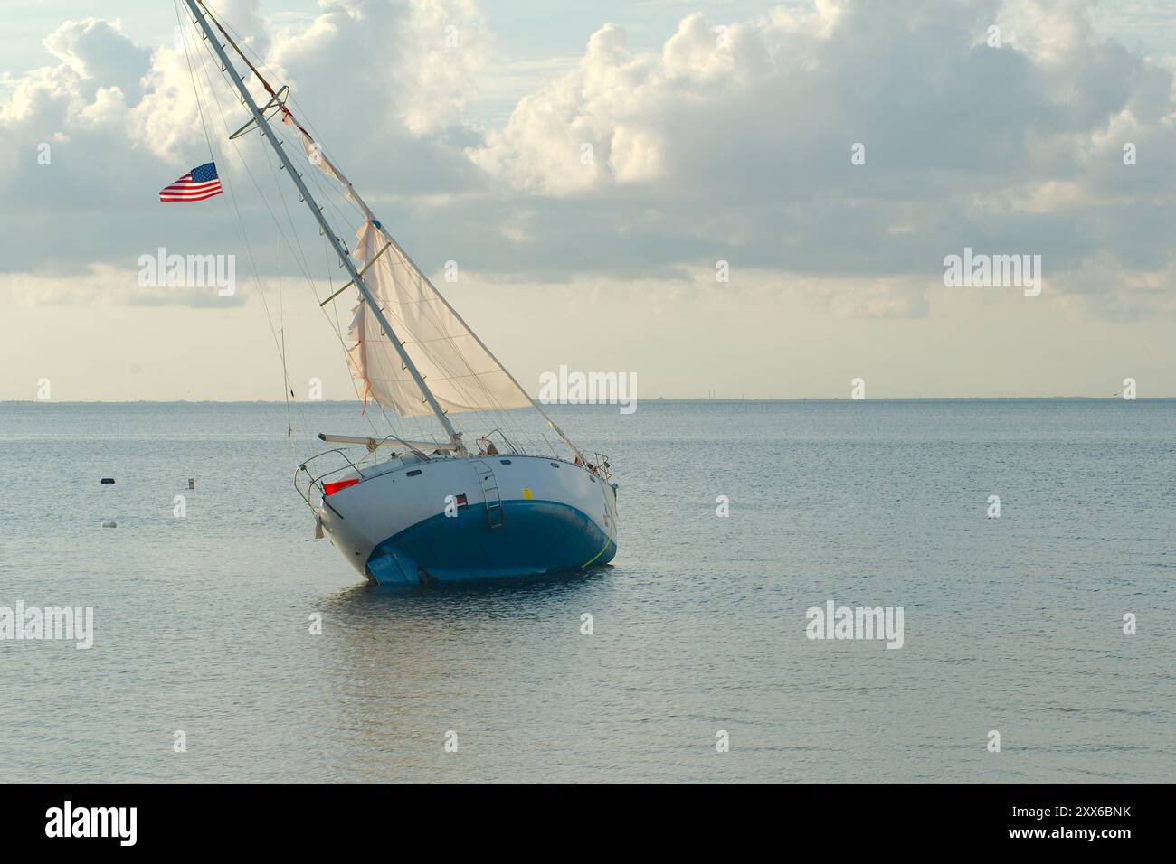 Vue large à l'est depuis North Shore Park sur Tampa Bay. Voilier bleu et blanc endommagé penché vers la droite avec voiles blanches et drapeau ondulant. Eau calme Banque D'Images