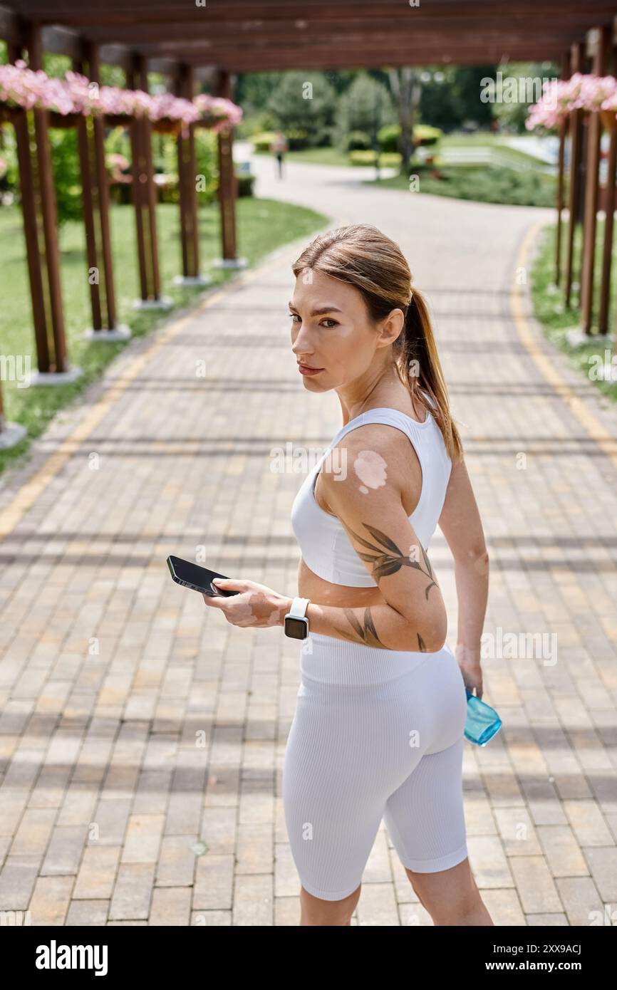 Une jeune femme atteinte de vitiligo porte des vêtements de sport blancs et une smartwatch alors qu'elle traverse un parc, tenant un téléphone et une bouteille d'eau. Banque D'Images