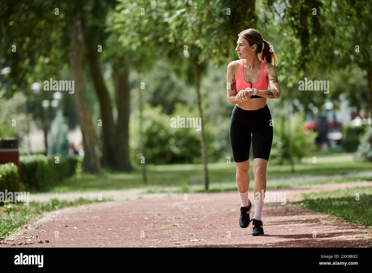 Une jeune femme atteinte de vitiligo vérifie sa montre en marchant sur un chemin de parc, portant un crop top rose et un short noir. Banque D'Images