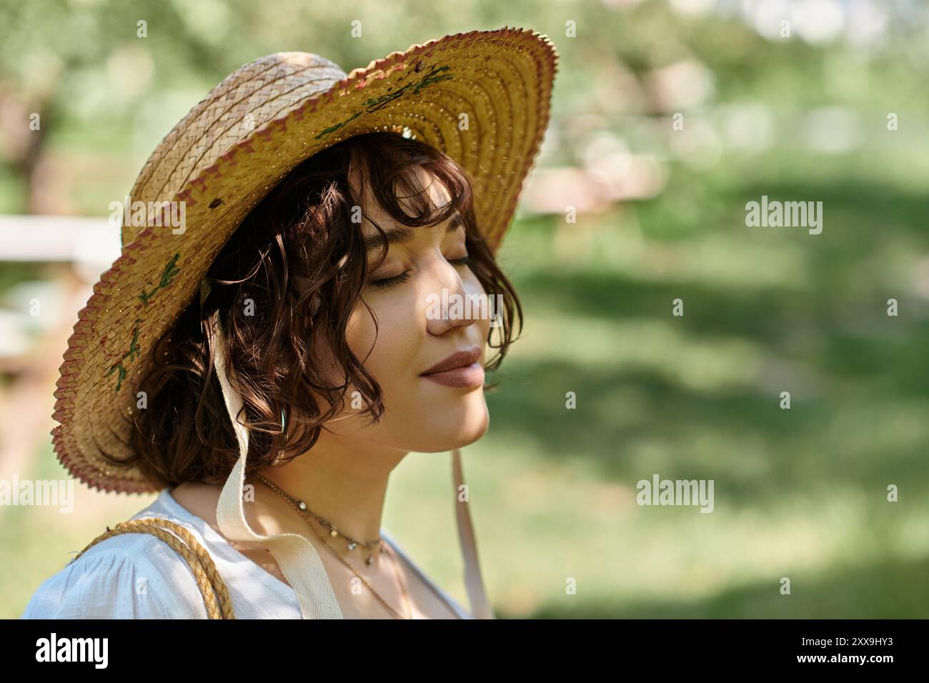 Une jeune femme aux cheveux bruns dans un chapeau de paille et un chemisier blanc se détend dans un jardin ensoleillé, les yeux fermés dans une contemplation paisible. Banque D'Images