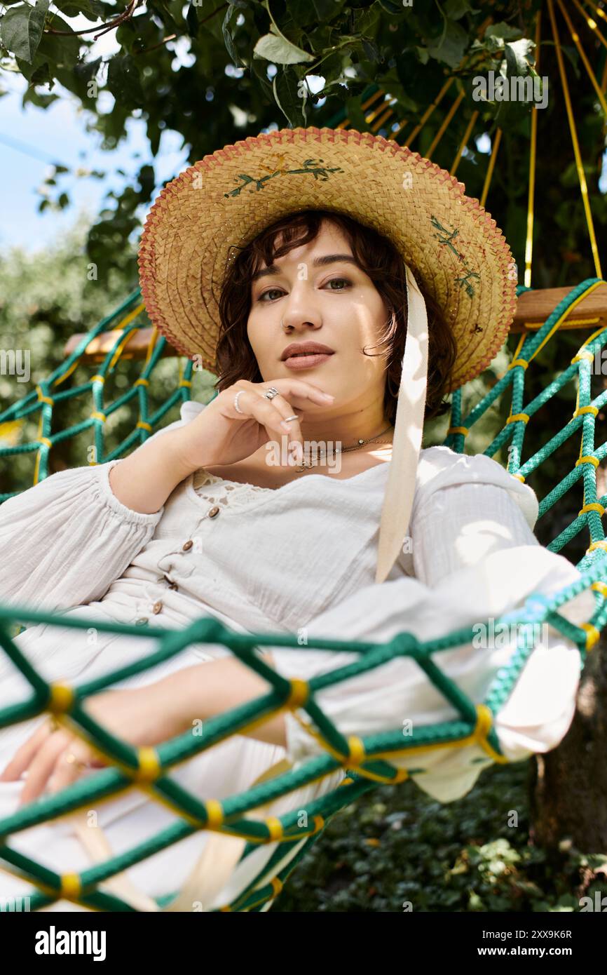 Une jeune femme en robe blanche et chapeau de paille se détend dans un hamac entouré de verdure. Banque D'Images