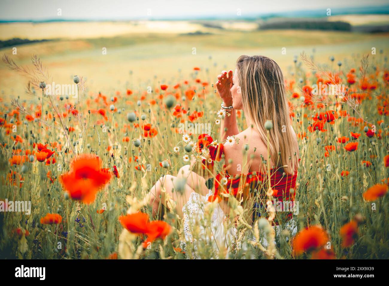 Jeune femme blonde en robe rouge et blanche est assise parmi les fleurs dans un champ de coquelicots rouges Banque D'Images