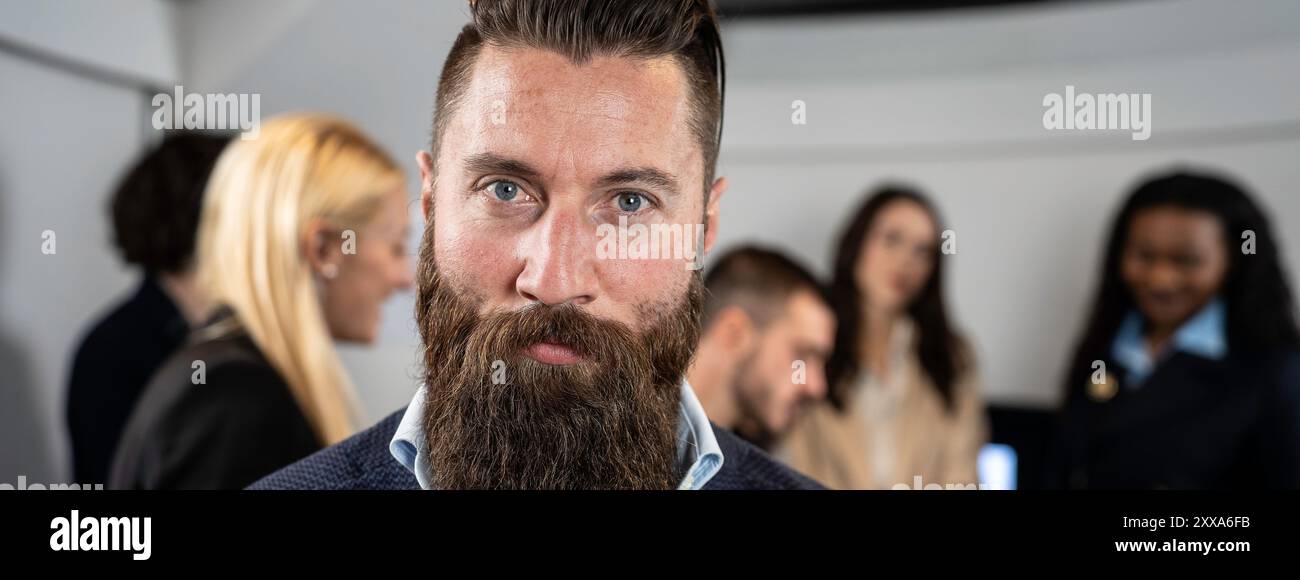 Bannière horizontale avec homme d'affaires confiant avec une expression sérieuse debout dans un bureau moderne, avec des collègues collaborant en arrière-plan. Banque D'Images