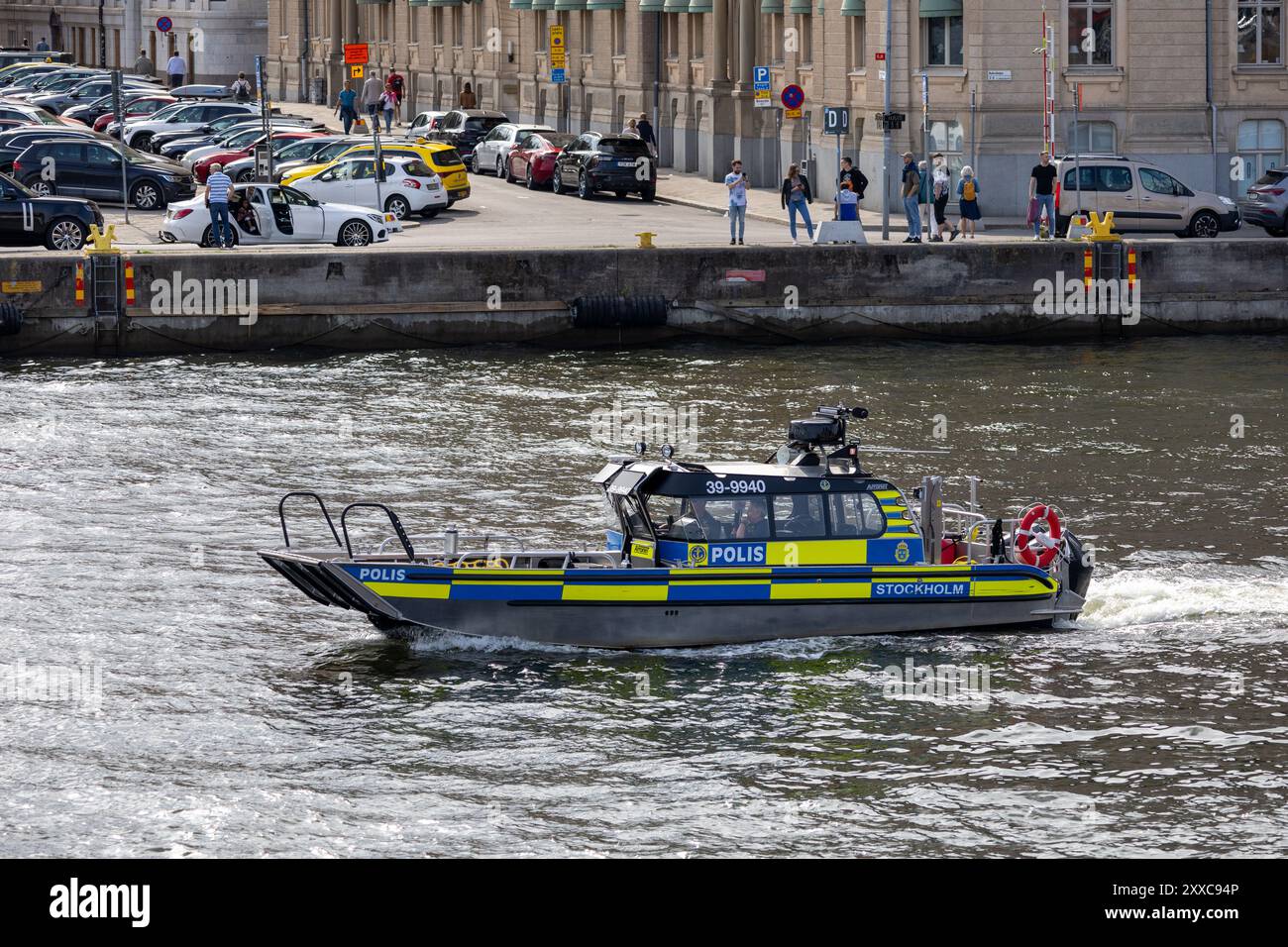 Un bateau de police naviguant sur une rivière avec un paysage urbain en arrière-plan. Les gens sont vus le long du front de mer, et diverses voitures sont garées à proximité. Banque D'Images