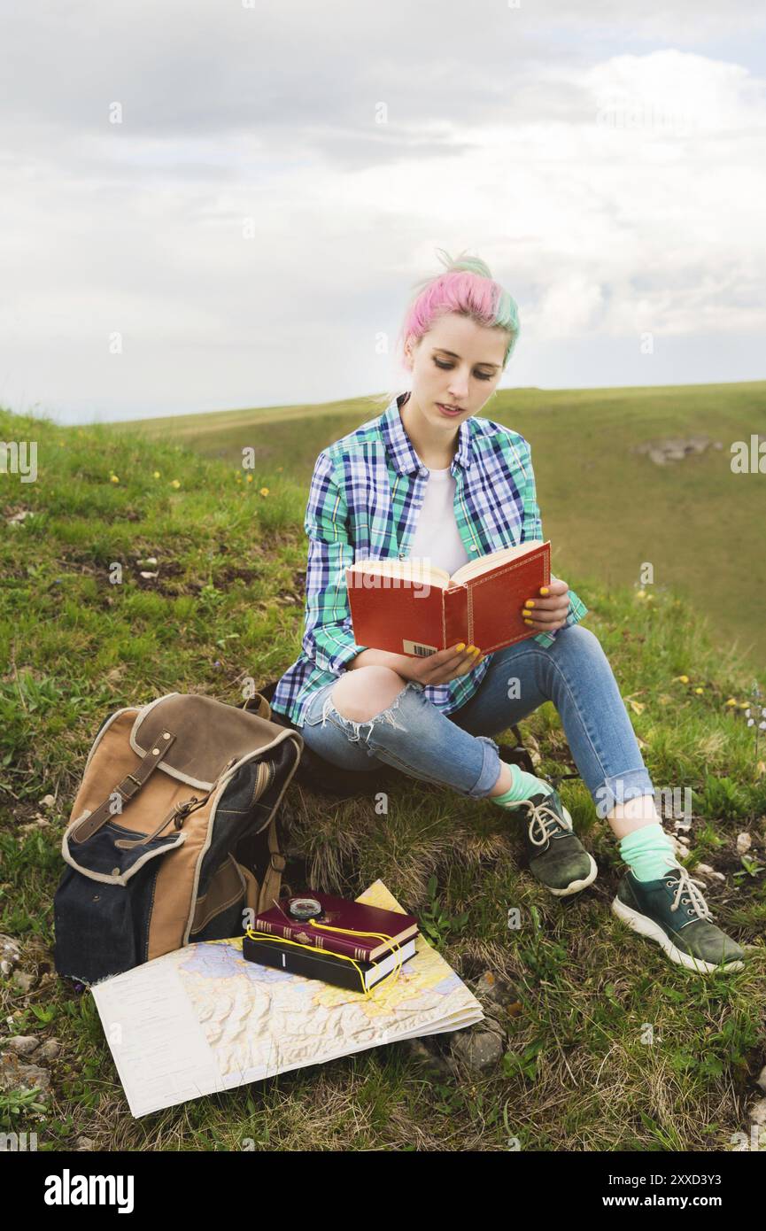 Une fille voyageuse est assise dans les montagnes sur l'herbe et lit un livre sur le fond de montagnes épiques. Le concept de lecture pendant le repos et vacati Banque D'Images