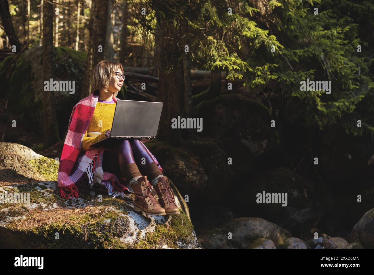 Un portrait tonique d'une jeune fille hipster freelance souriante avec des lunettes habillées dans une couverture avec un ordinateur portable agenouillé assis sur un rocher dans un conifère pour Banque D'Images