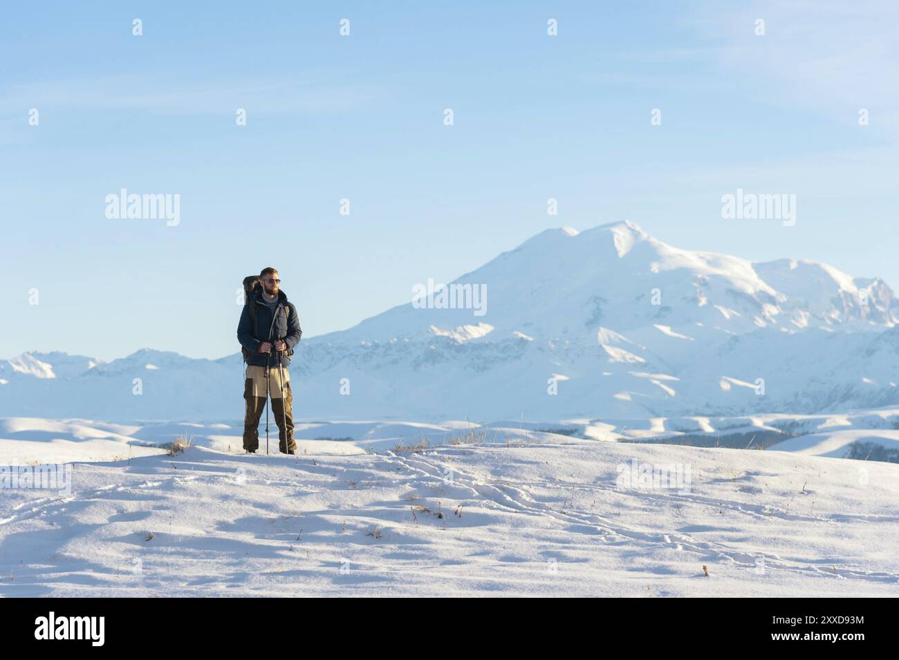 Un voyageur avec un grand sac à dos sur ses épaules se tient sur une colline enneigée contre le ciel bleu et le volcan Elbrus endormi entouré par le M. Banque D'Images