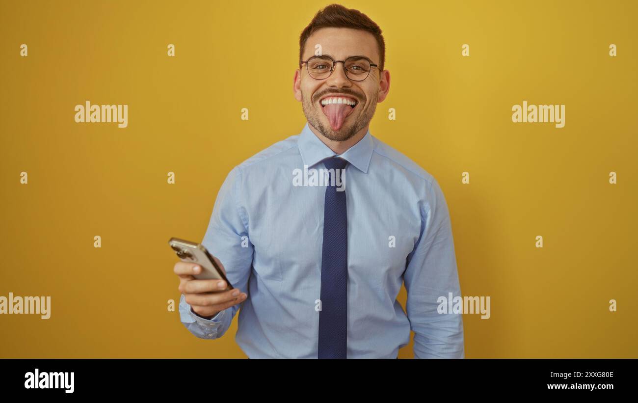 Un beau jeune homme hispanique avec une barbe et des lunettes sort sa langue tout en tenant un téléphone, isolé sur un fond jaune vif. Banque D'Images