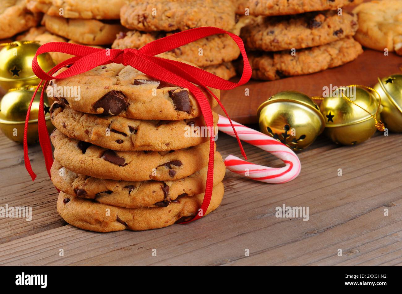 Une pile de biscuits aux pépites de chocolat des fêtes noués avec un ruban rouge devant des cloches de jingle, une canne de bonbons, et un plateau de friandises assorties. Horizo Banque D'Images