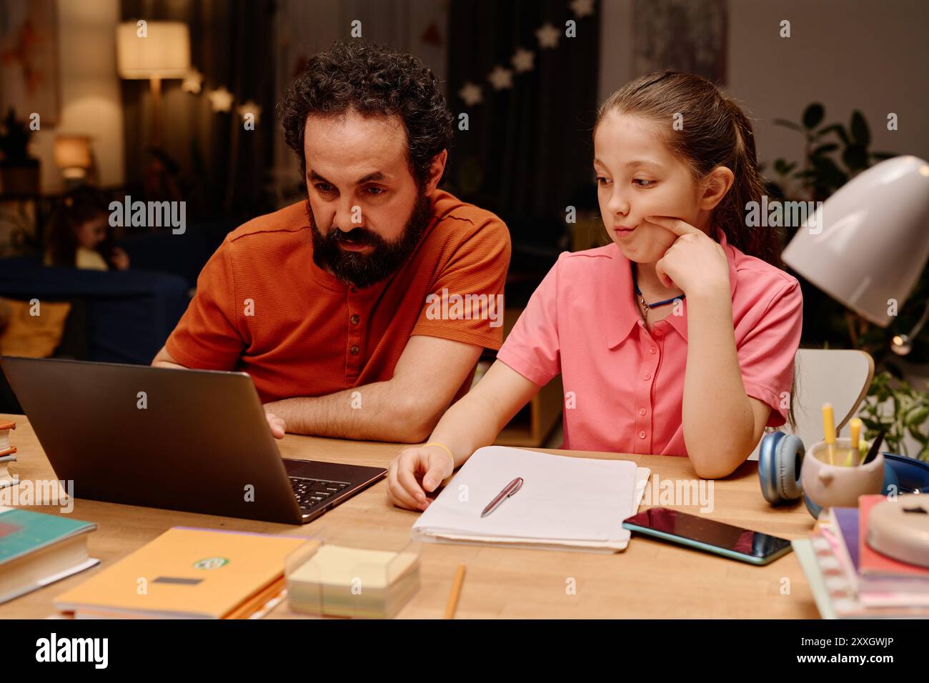 Homme barbu aidant la jeune fille avec les devoirs, tous deux concentrés sur l'écran d'ordinateur portable, divers articles de papeterie sur le bureau autour d'eux. Père fournissant de l'aide à la fille dans un environnement familial confortable Banque D'Images