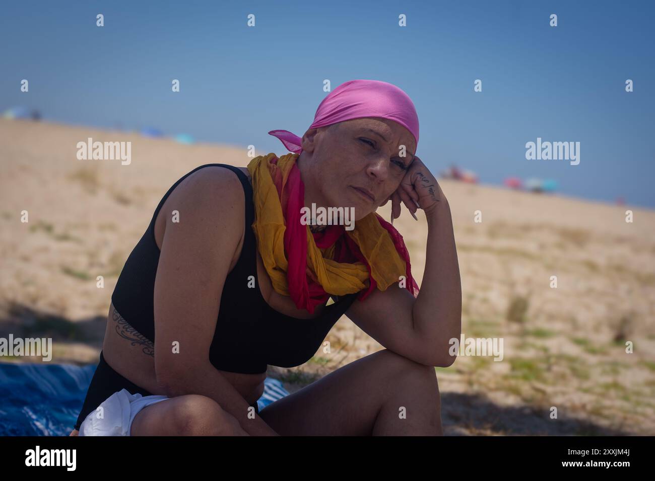 Femme d'âge moyen atteinte d'un cancer assise réfléchie sur une couverture de plage, regardant au loin Banque D'Images