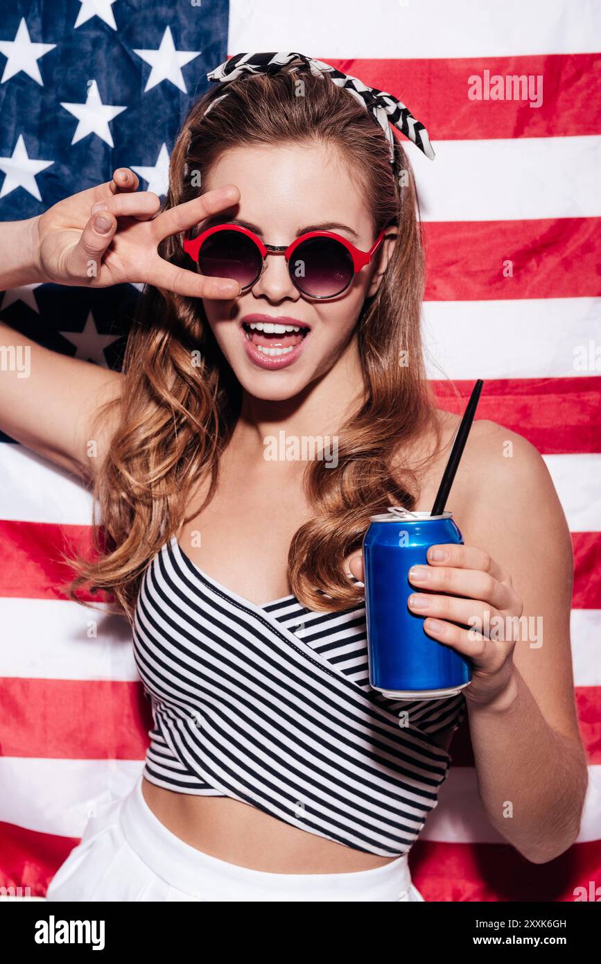 La liberté est dans ses veines. Belle jeune femme donnant la paix et tenant une canette tout en se tenant debout contre le drapeau national américain Banque D'Images