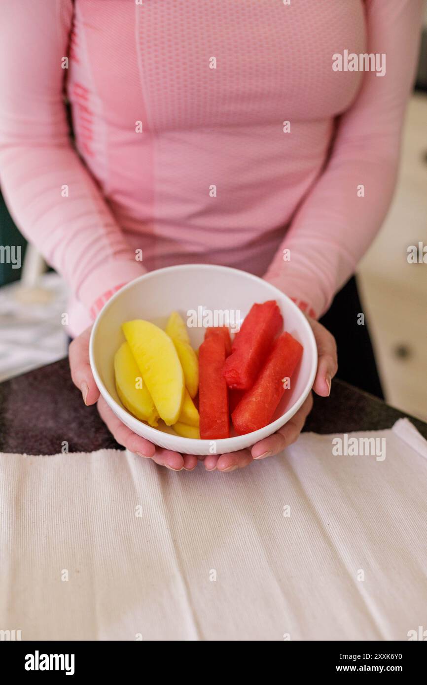 Une femme en équipement de fitness tient un bol de fruti contenant des doigts de mangue et de pastèque pour fournir une collation nutritive et énergisante Banque D'Images