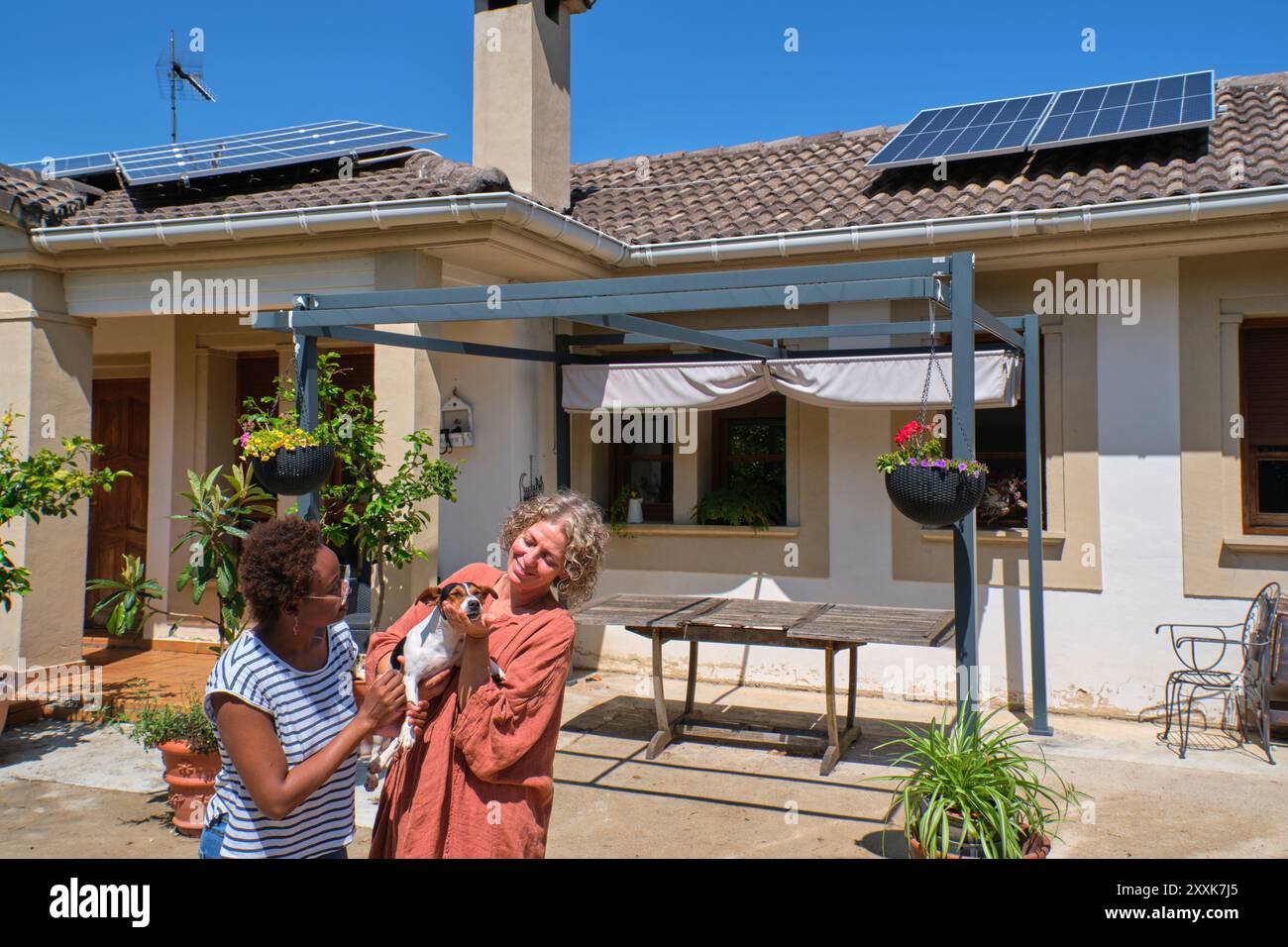 Deux femmes se tiennent joyeusement ensemble dans une cour ensoleillée, tenant un petit chien. Le cadre est paisible, avec des plantes en pot et des panneaux solaires visibles Banque D'Images