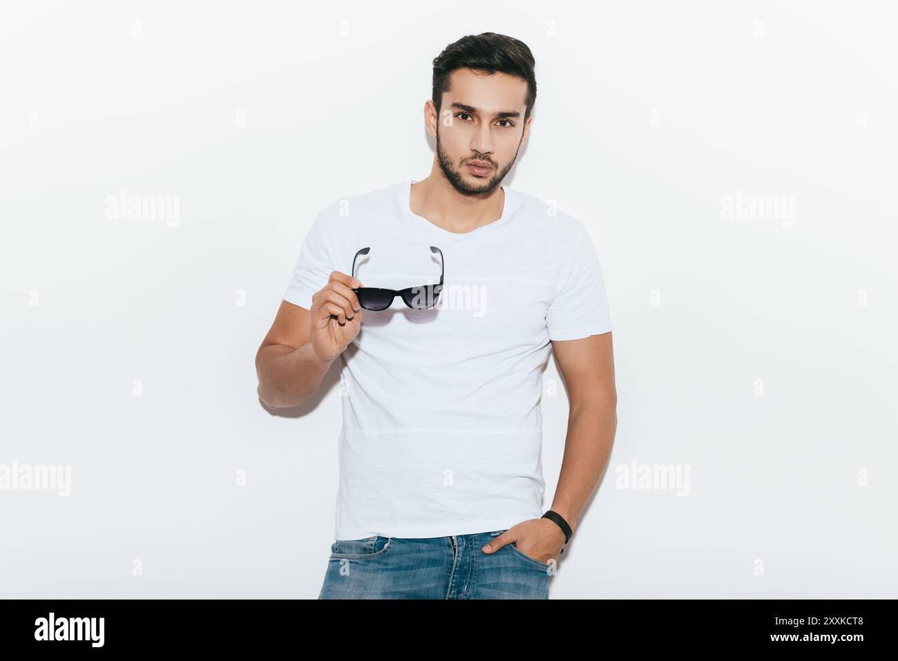 Charmant beau. Beau jeune homme indien portant des lunettes de soleil et regardant la caméra tout en se tenant debout sur fond blanc Banque D'Images