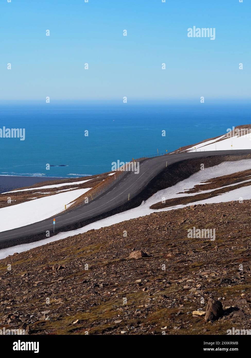 Une route sinueuse courbe à travers un terrain de haute altitude avec des plaques de neige, offrant une vue panoramique sur l'immense océan bleu rencontrant l'horizon sous-jacent Banque D'Images
