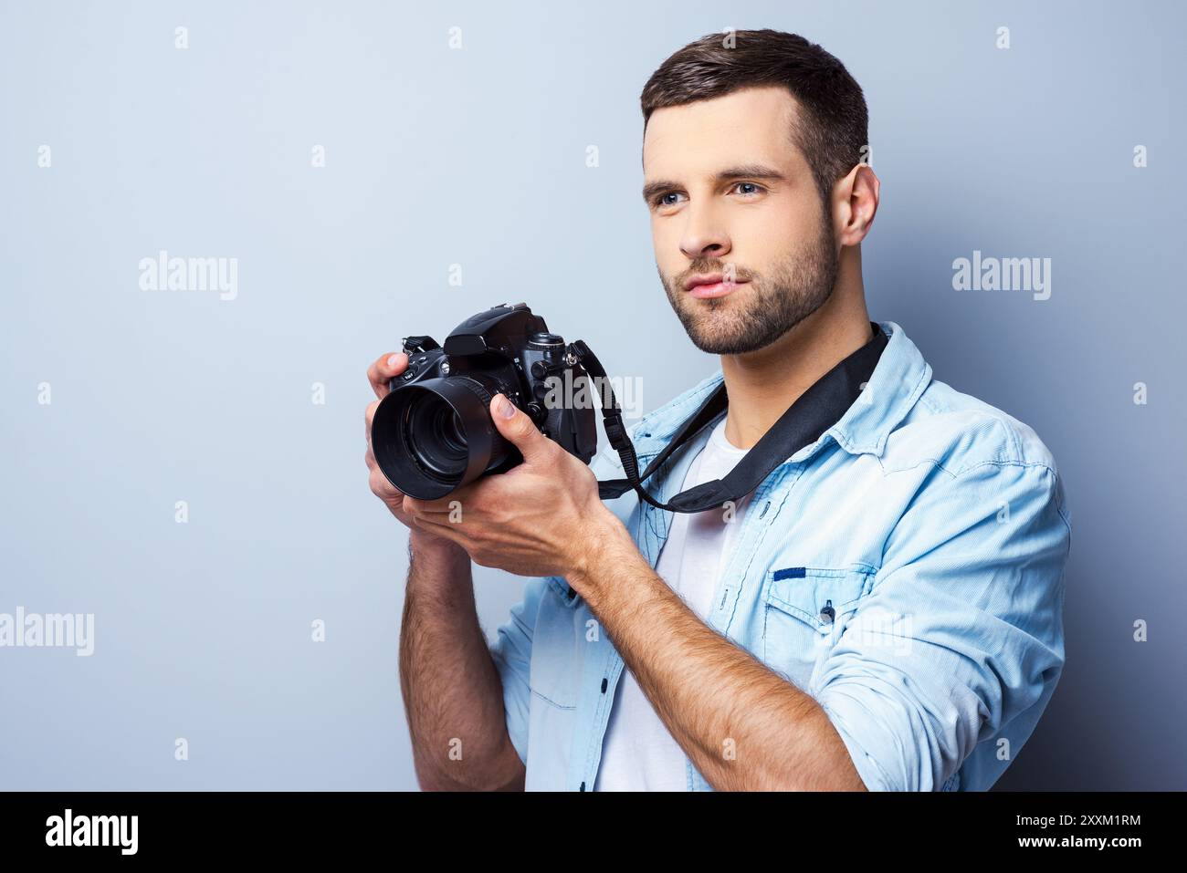 Je serai un excellent tir. Beau jeune homme tenant un appareil photo numérique et regardant loin tout en se tenant debout sur fond gris Banque D'Images