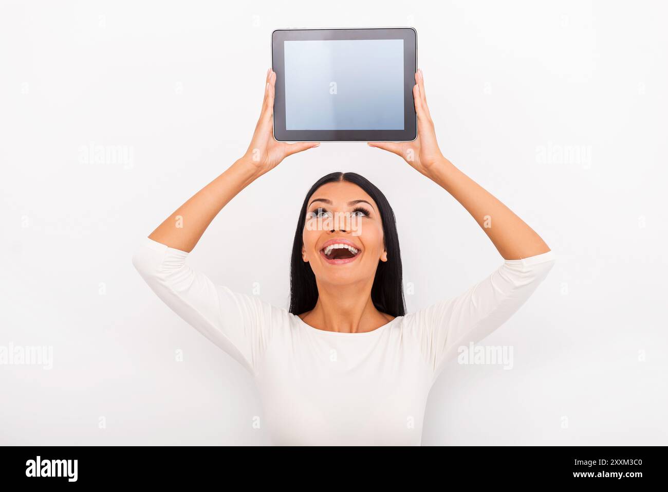 Elle aime les gadgets intelligents. Jeune femme joyeuse tenant une tablette numérique au-dessus de sa tête tout en se tenant debout sur fond blanc Banque D'Images