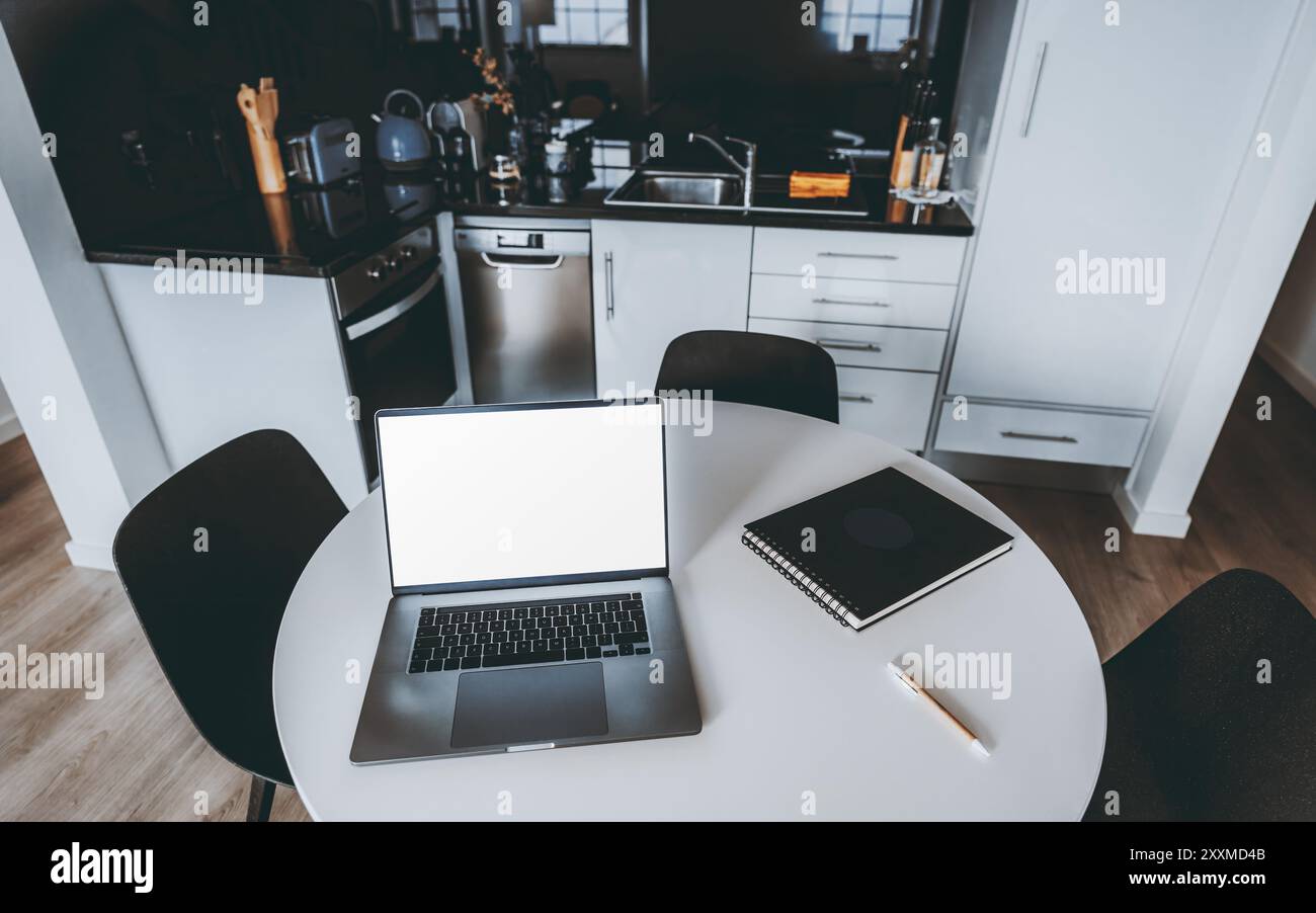 Une configuration de cuisine moderne avec une table ronde blanche, ordinateur portable ouvert avec maquette d'écran, ordinateur portable et stylo. L'arrière-plan présente des appareils de cuisine élégants a Banque D'Images