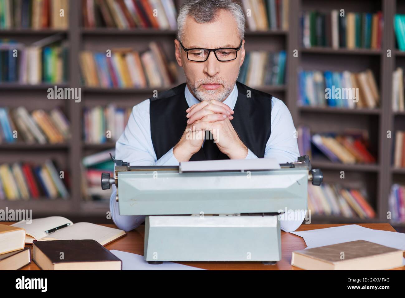En attendant l'inspiration. Confiant cheveux gris homme senior en vêtements de forme assis à la machine à écrire et regardant concentré avec étagère dans le backgro Banque D'Images