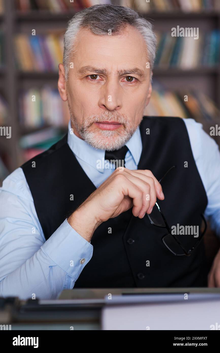 Auteur confiant. Homme senior de cheveux gris confiant en vêtements de forme assis à la machine à écrire et regardant la caméra avec étagère à l'arrière-plan Banque D'Images
