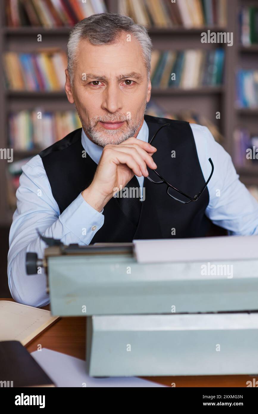 Juste inspiré. Homme senior de cheveux gris confiant en vêtements de forme assis à la machine à écrire et regardant la caméra avec étagère à l'arrière-plan Banque D'Images