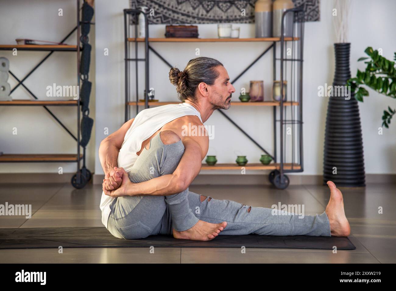 Vue latérale d'un athlète masculin adulte aux pieds nus fort en vêtements de sport regardant loin tout en étant assis sur le tapis et en faisant la pose de yoga Marichyasana III dans le hall pendant da Banque D'Images