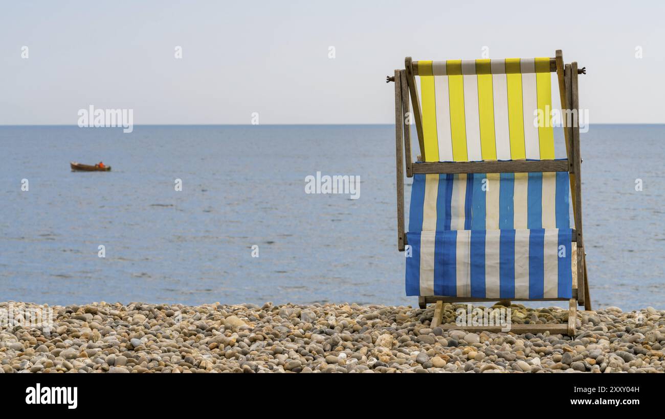 Chaise Empty Deck sur la plage de galets à Beer, Devon, Royaume-Uni, avec un bateau de pêche dans la baie de Seaton au canal britannique Banque D'Images